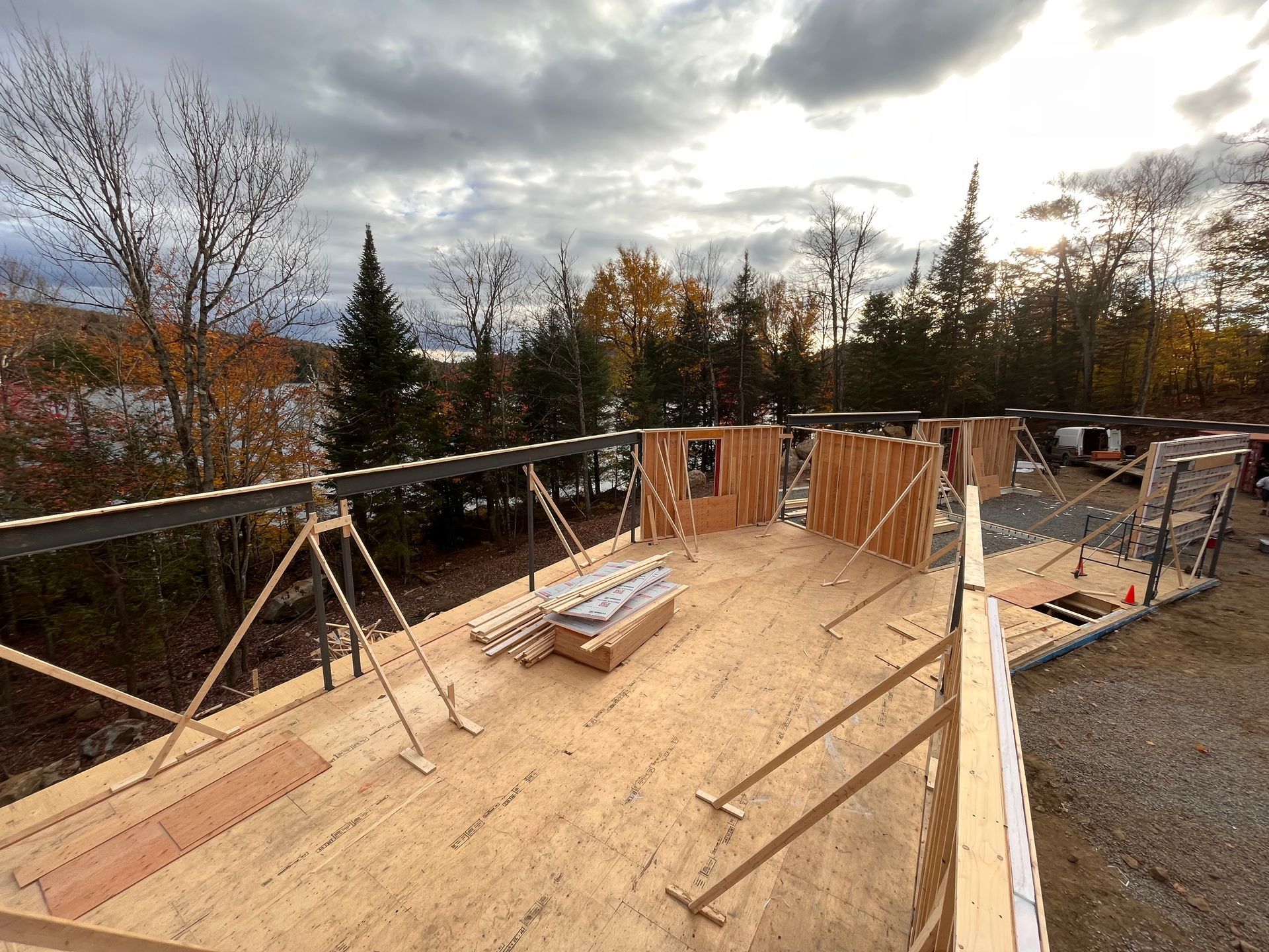 Chantier : charpente en bois avec poutres apparentes, donnant sur un lac et des arbres sous un ciel nuageux.