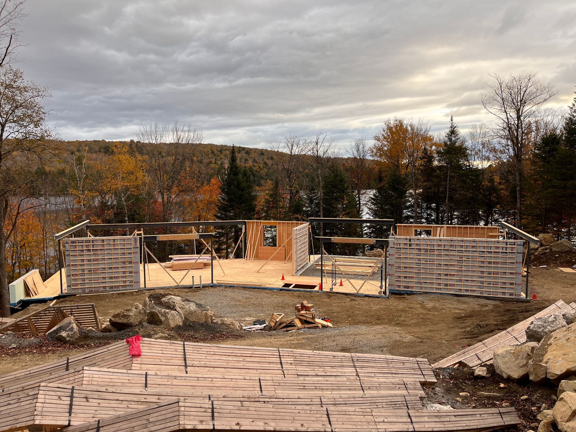 Chantier de construction avec des charpentes en bois et des palettes sur un terrain nivelé, avec une vue pittoresque en arrière-plan sous un ciel nuageux.