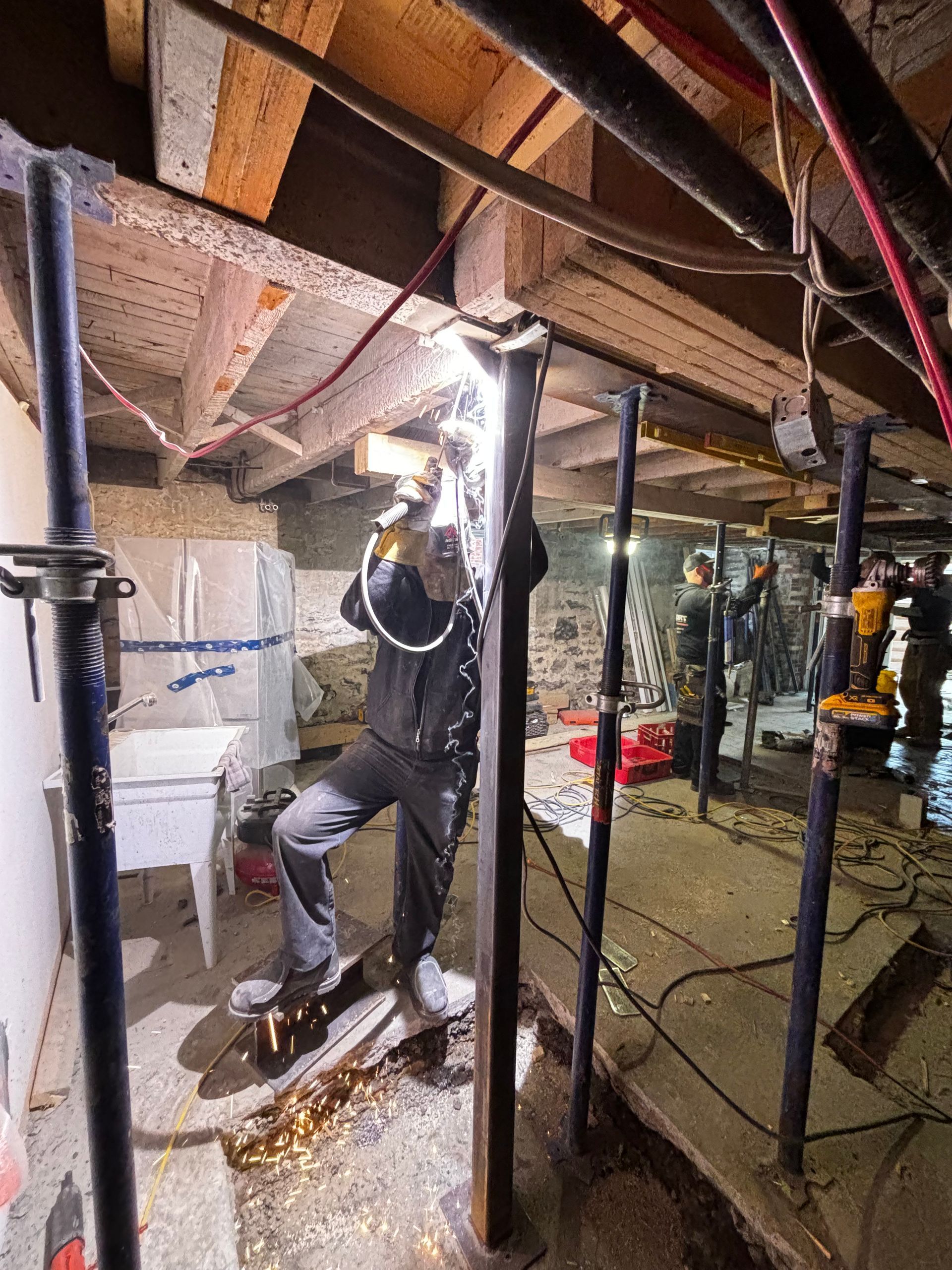 Un ouvrier soude des supports métalliques dans un sous-sol faiblement éclairé. Des étincelles jaillissent tandis qu'il travaille près des poutres et des colonnes de soutien apparentes.