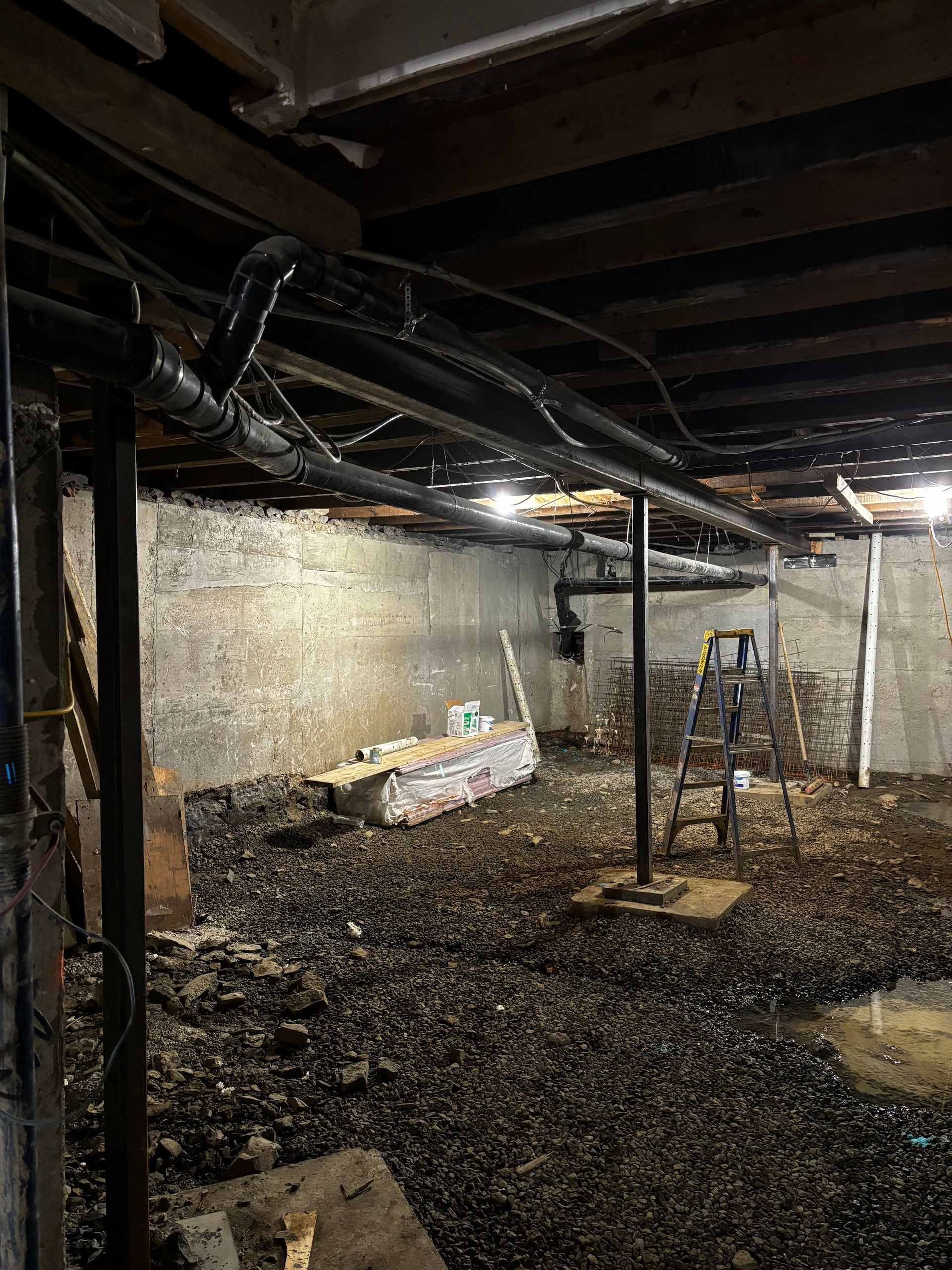 Sous-sol avec tuyaux apparents, murs en béton, sol en gravier et matériaux de construction.