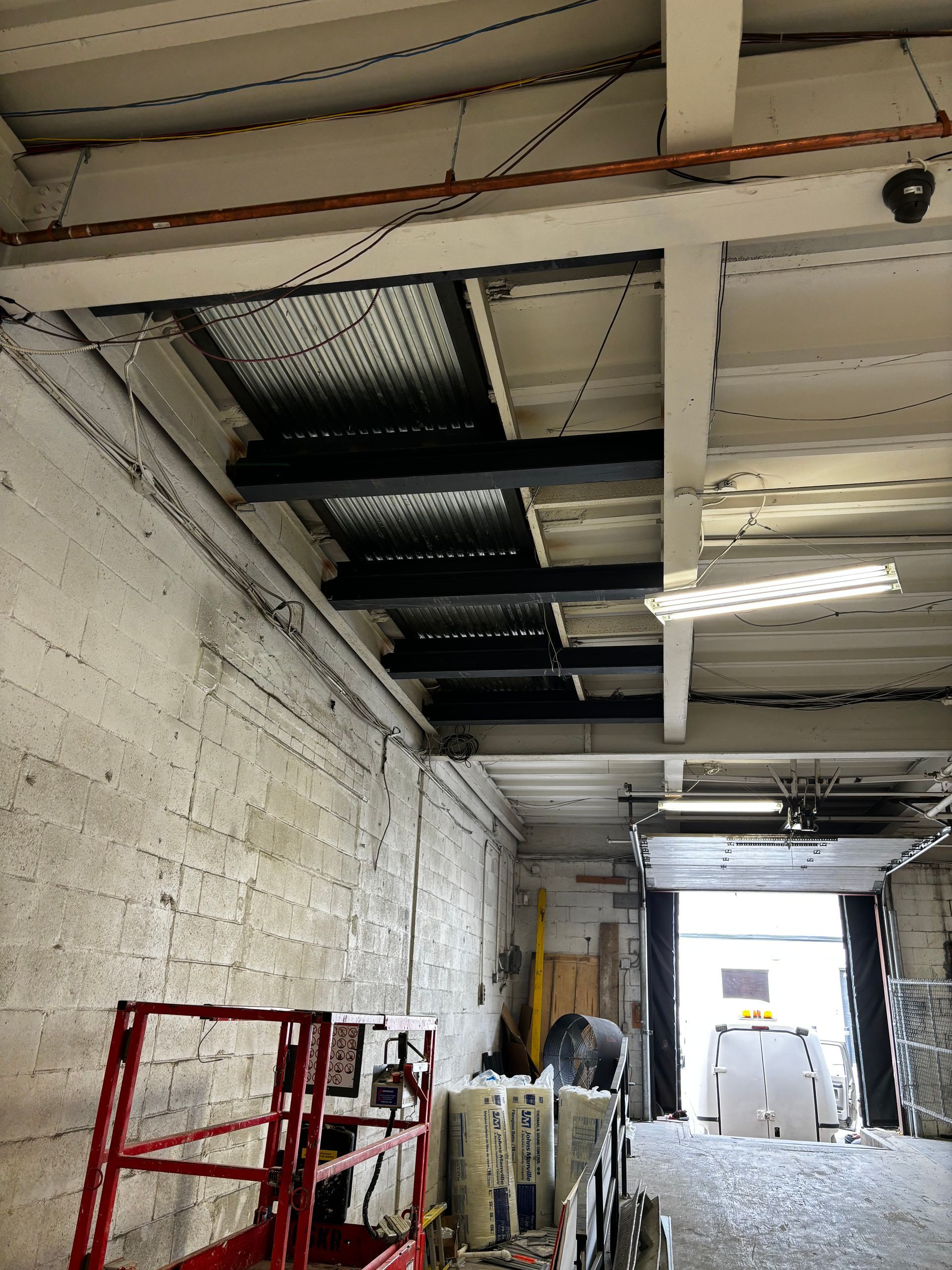 Intérieur d'un entrepôt avec un haut plafond, des poutres apparentes et une porte ouverte donnant sur l'extérieur.