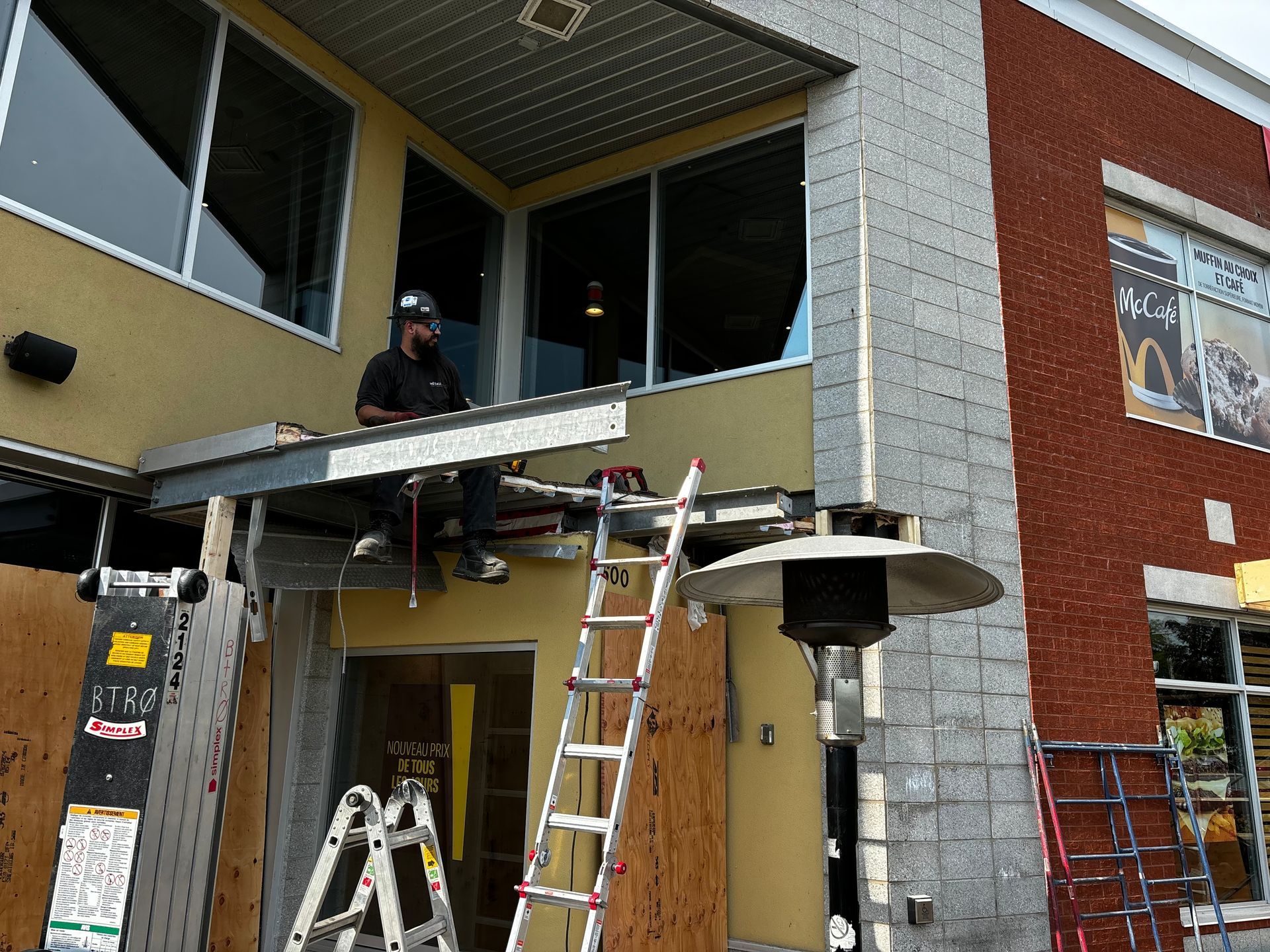 Un ouvrier du bâtiment, sur une plateforme, installe un auvent au-dessus de l'entrée d'un bâtiment commercial ; outils et échelle sont visibles.