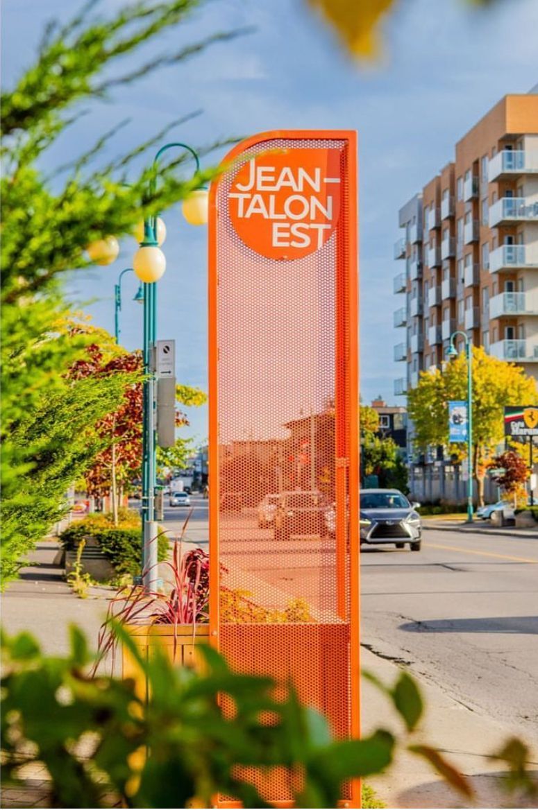 Panneau orange Jean-Talon Est sur une rue de la ville, visible à travers le feuillage vert.