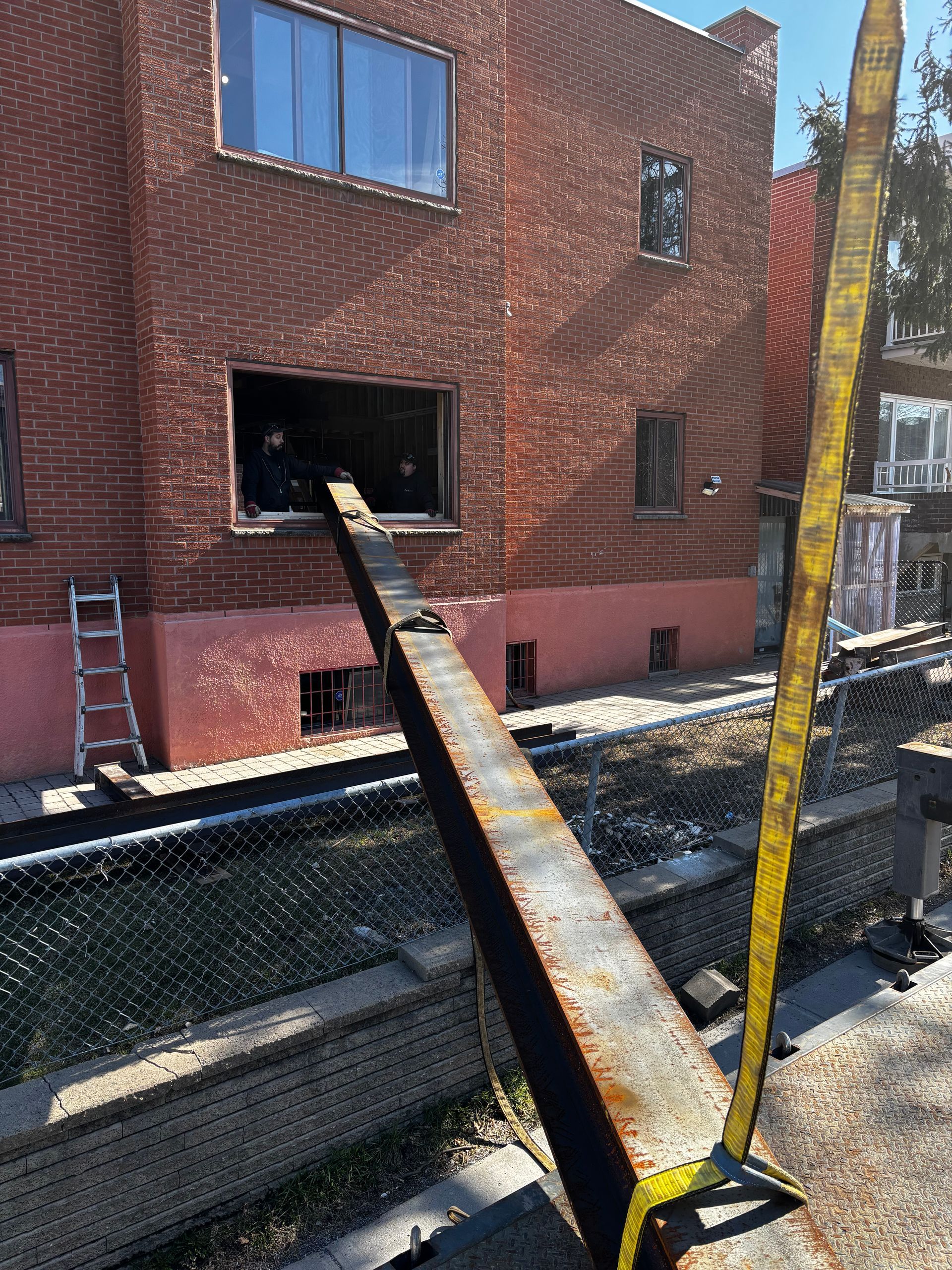Une poutre d'acier fixée à la fenêtre d'un bâtiment en briques rouges est maintenue par une sangle jaune.