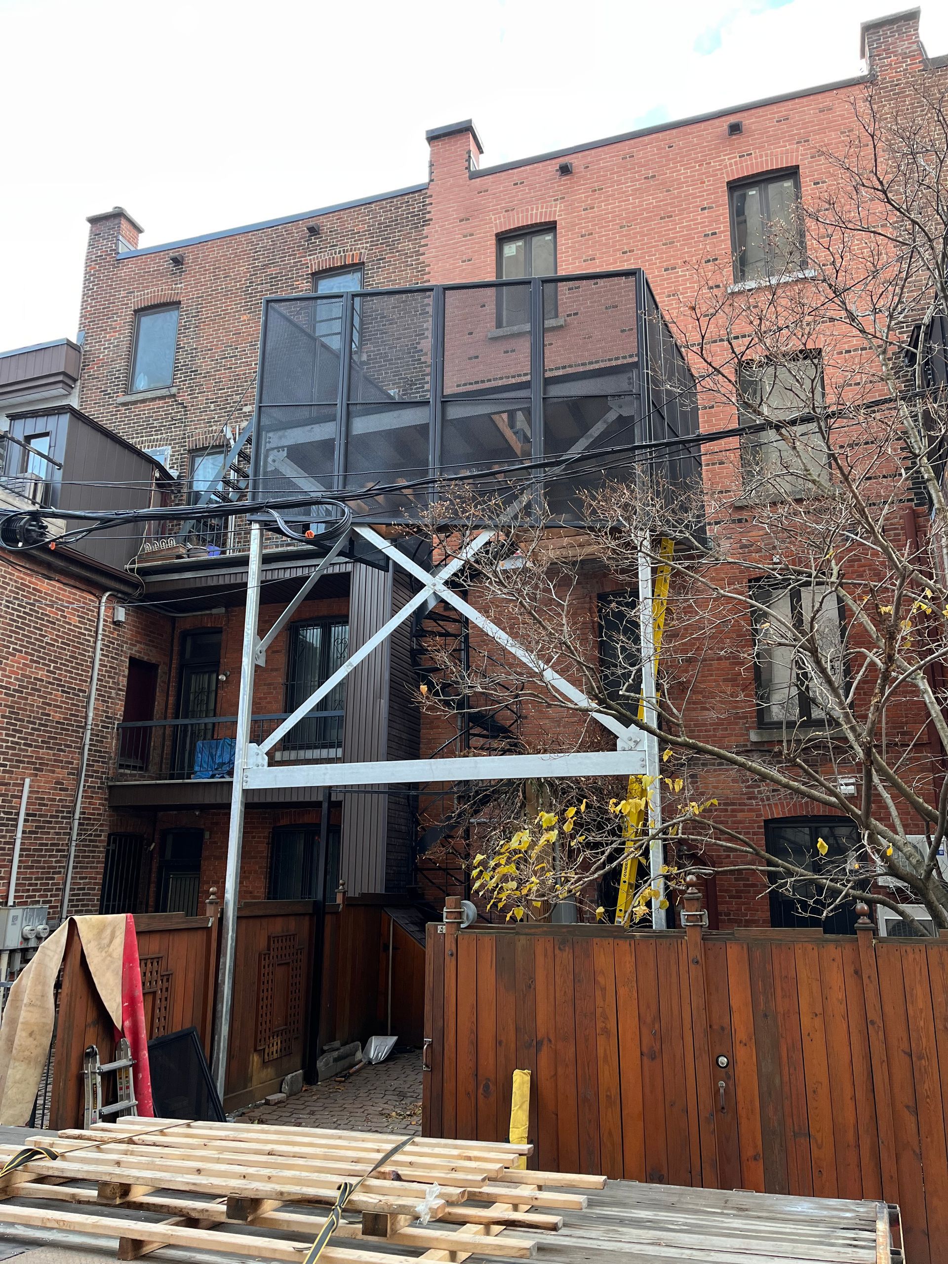 Échafaudage adossé à un bâtiment en briques rouges, soutenant une structure en treillis noir. Clôture en bois au premier plan.