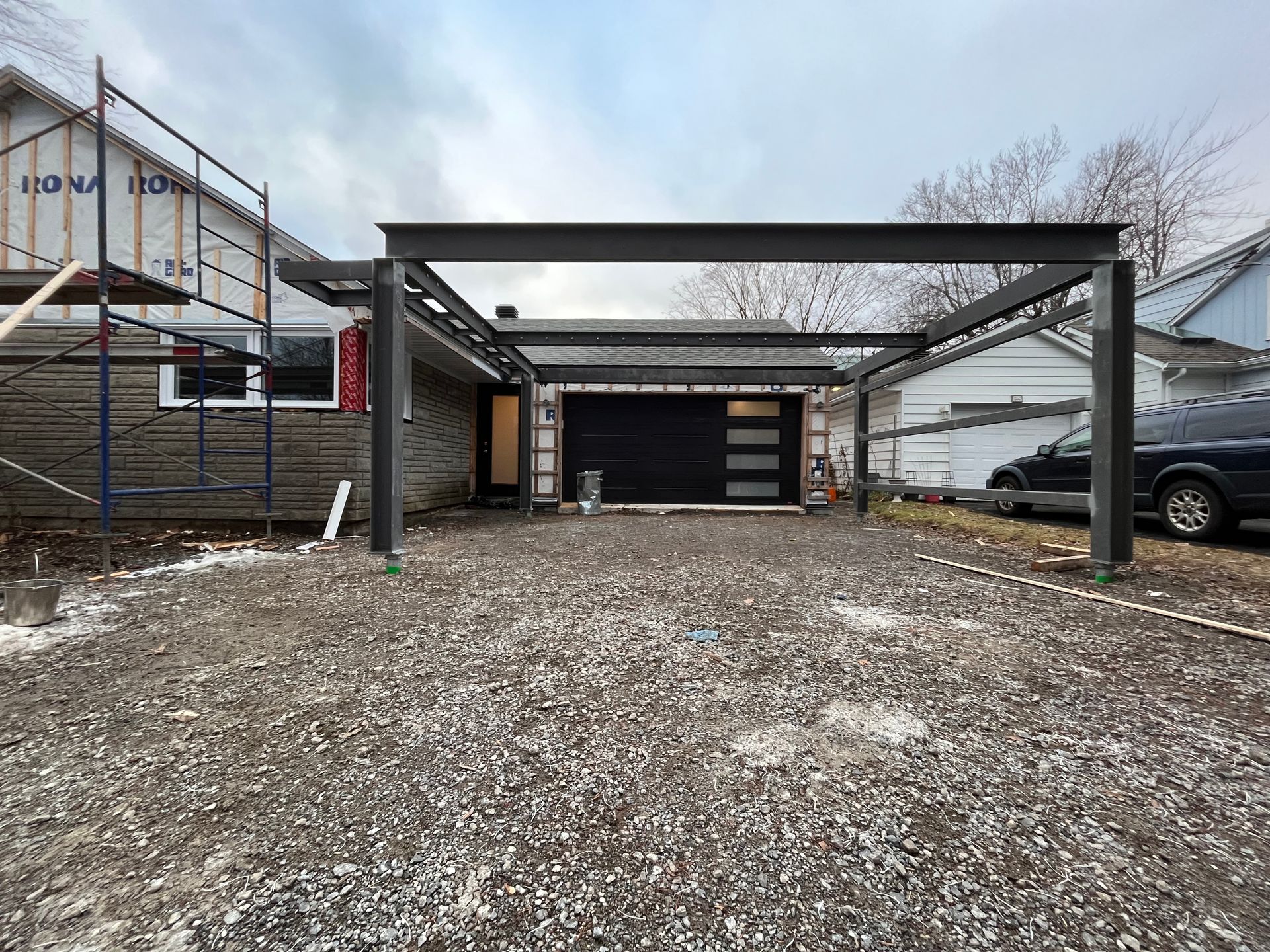 Allée avec un abri de voiture métallique au-dessus d'une porte de garage ; maison en construction à gauche, véhicules à droite.