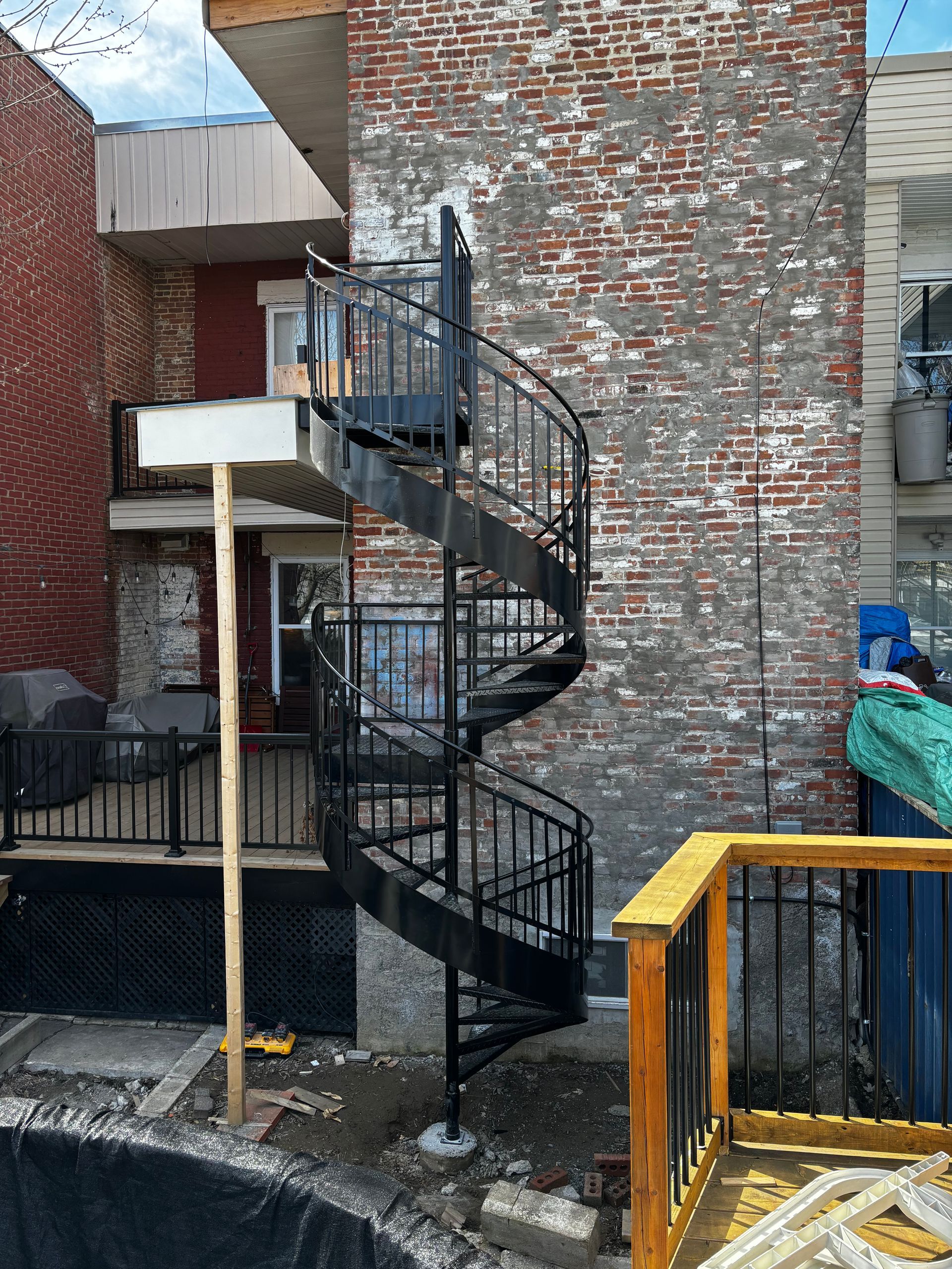 Un escalier en colimaçon noir se détache sur un bâtiment en briques patinées. Une terrasse en bois se trouve au premier plan.