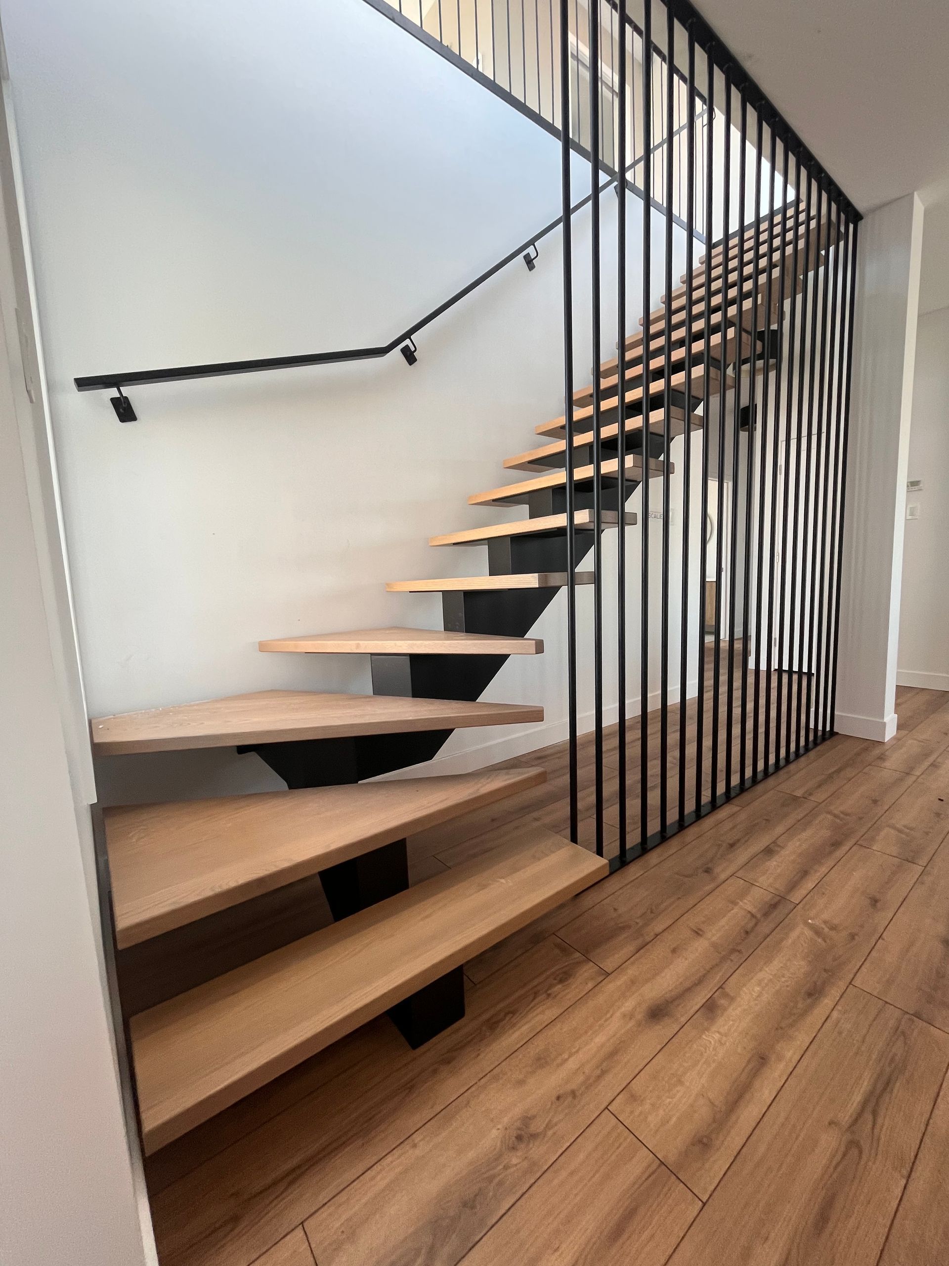 Escalier en bois avec supports et rampe en métal noir, adossé à un mur blanc.
