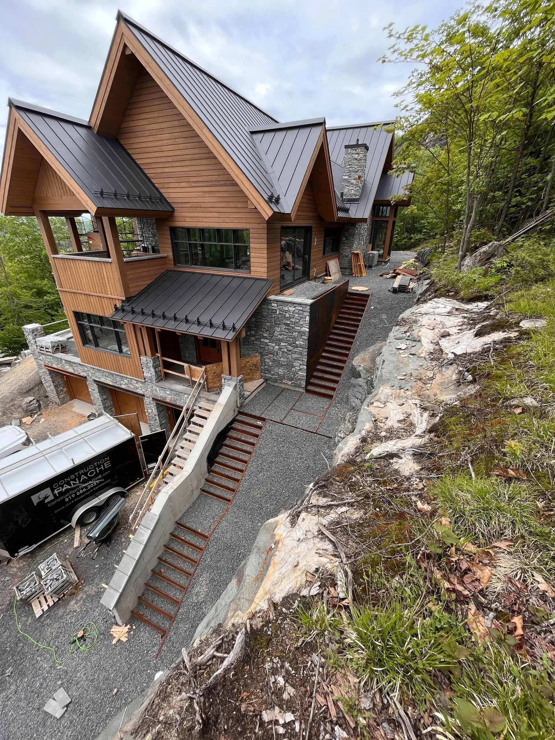 Maison en bois à plusieurs niveaux, construite à flanc de colline, avec des escaliers et des allées de gravier menant à l'entrée.