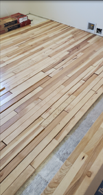 Unfinished light-colored wooden floor planks being installed in a room with white walls and exposed underlayment.