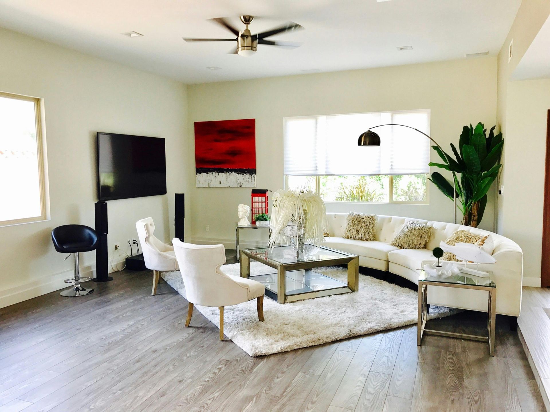 A modern living room with a white curved sofa, two white armchairs, a glass coffee table, and a large wall-mounted TV.