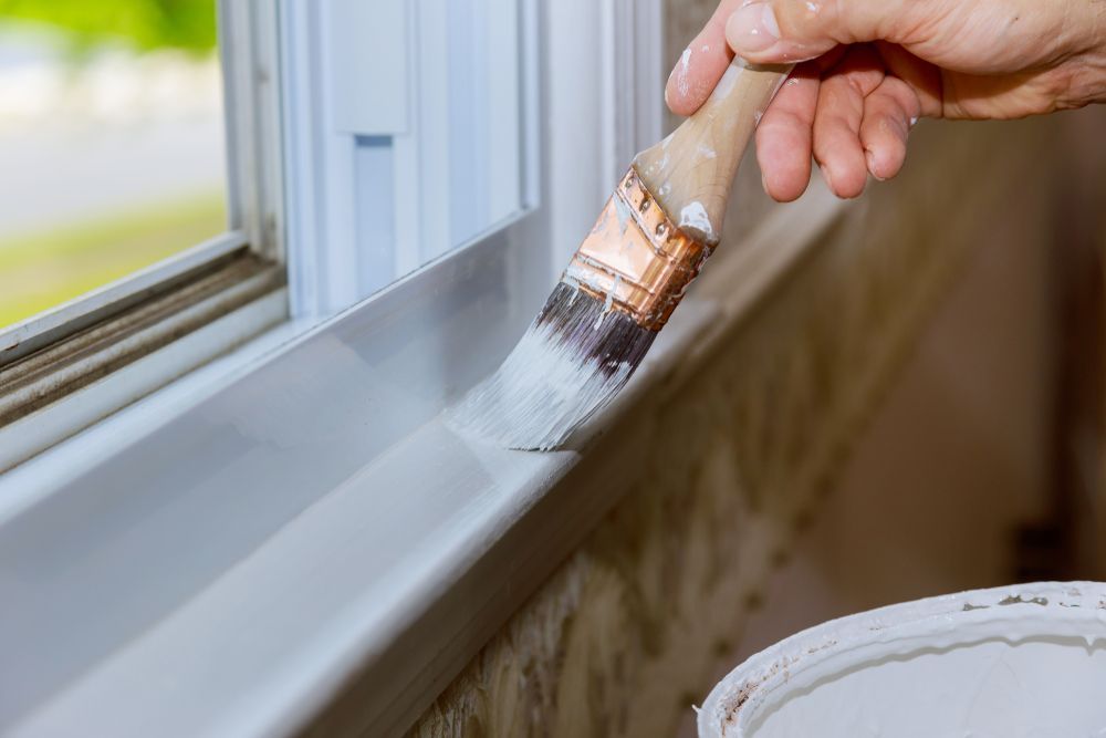 A close-up of a hand using a brush to paint a white window sill.