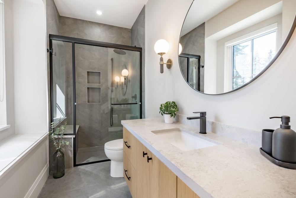 A modern bathroom with a light wood vanity, white countertop, round mirror, and a walk-in glass shower with grey tile.