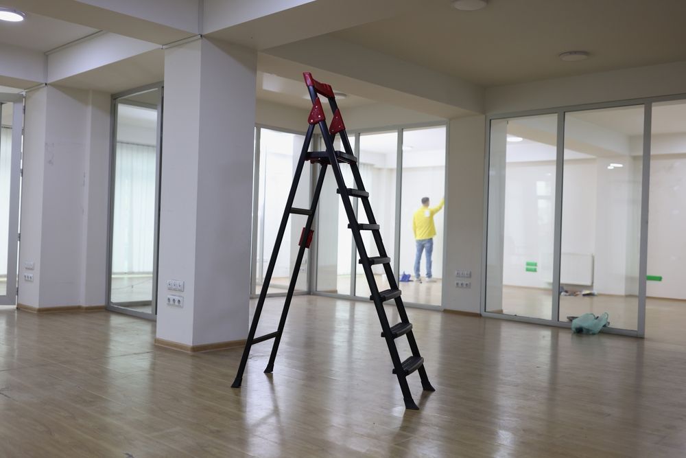 A black stepladder stands in the center of an empty office, with a person in a yellow jacket working in the background.