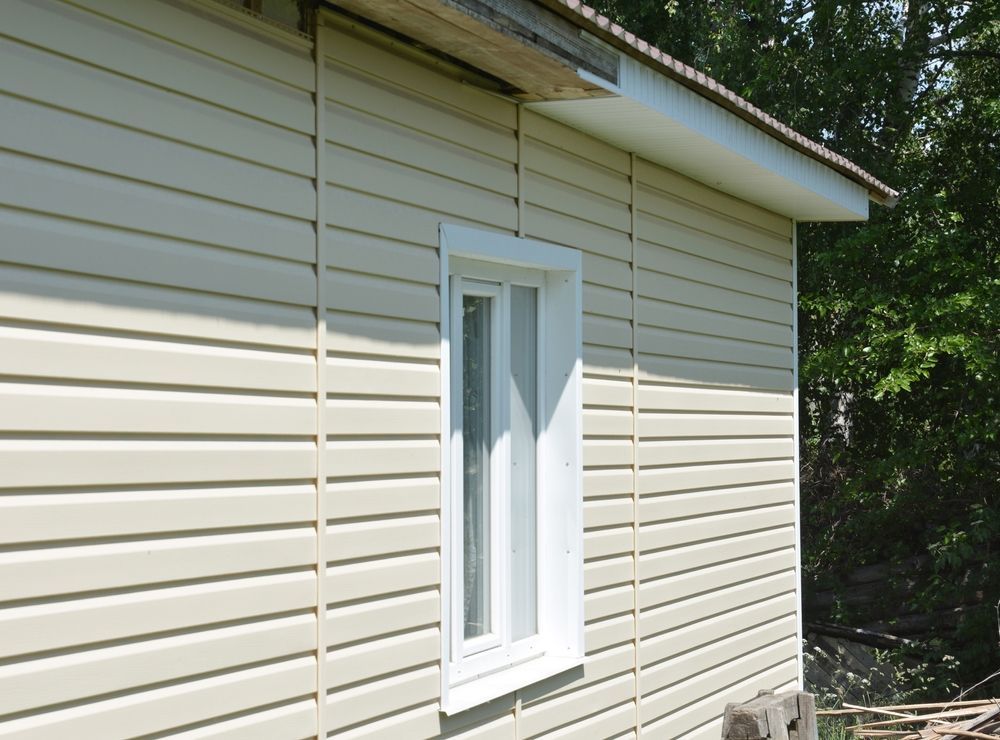 A corner of a house exterior clad in light beige horizontal vinyl siding with a white-framed window.