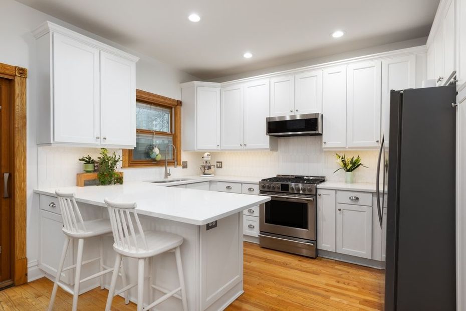 A bright kitchen with white cabinets, light countertops, two stools at a small island, and stainless steel appliances.