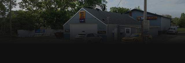 A blue garage-like building with a large garage door and a yellow sign on the roof. Trees surround it.