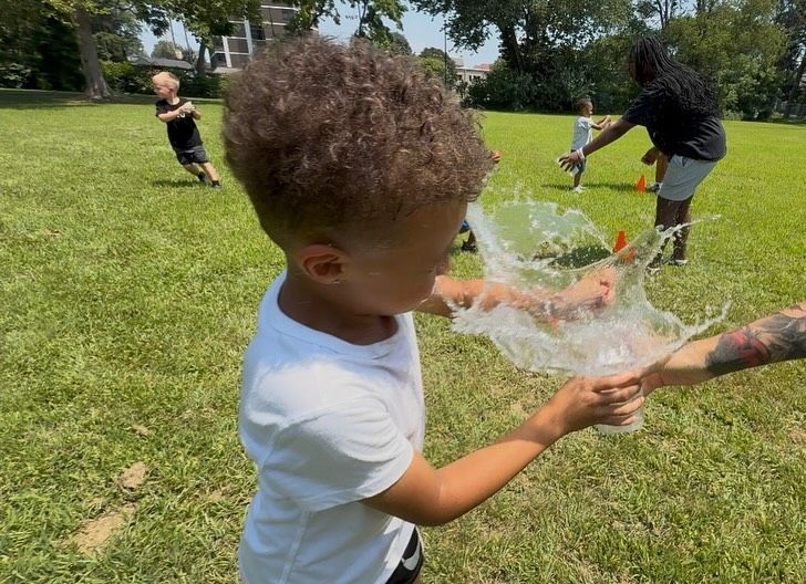 Boy with curly hair splashes water from a cup outdoors. People and a green field are in the background.
