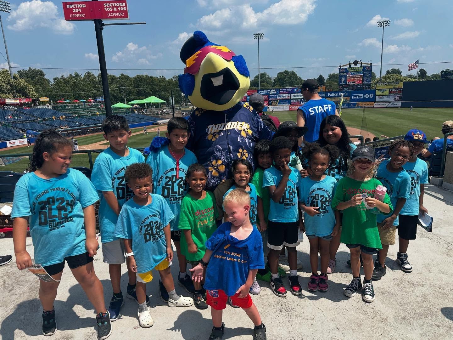 Children in blue and green shirts with mascot at baseball field.
