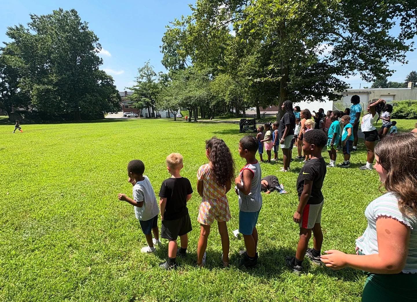 Children standing in a grassy field, watching a game. Green grass, blue sky, and trees in the background.