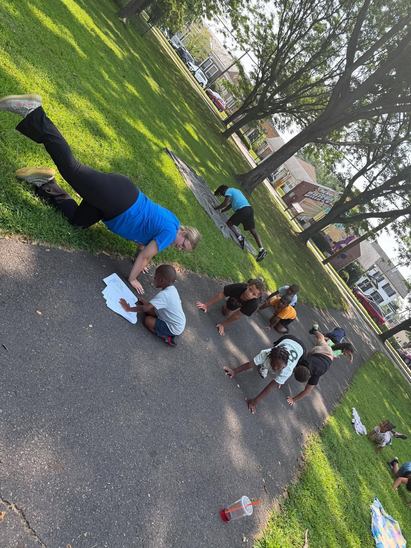 Group doing outdoor exercises, with instructor and child on pavement, green grass, trees and houses in background.
