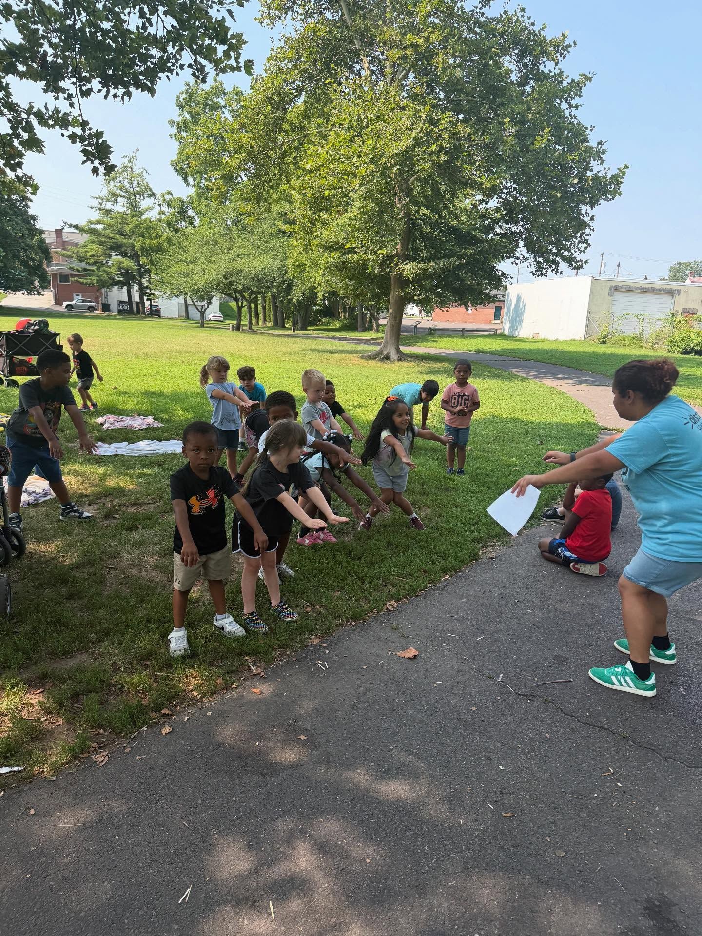 Children stretching in a park, led by an instructor holding papers on a sunny day.
