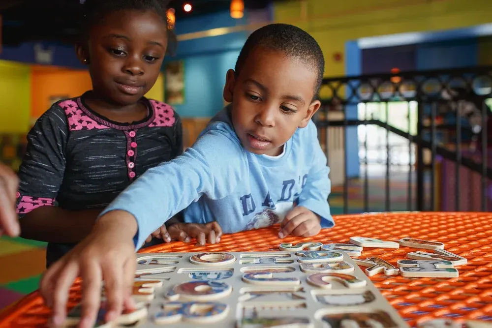 Two children working together to complete a wooden puzzle on an orange table inside a brightly colored play area.