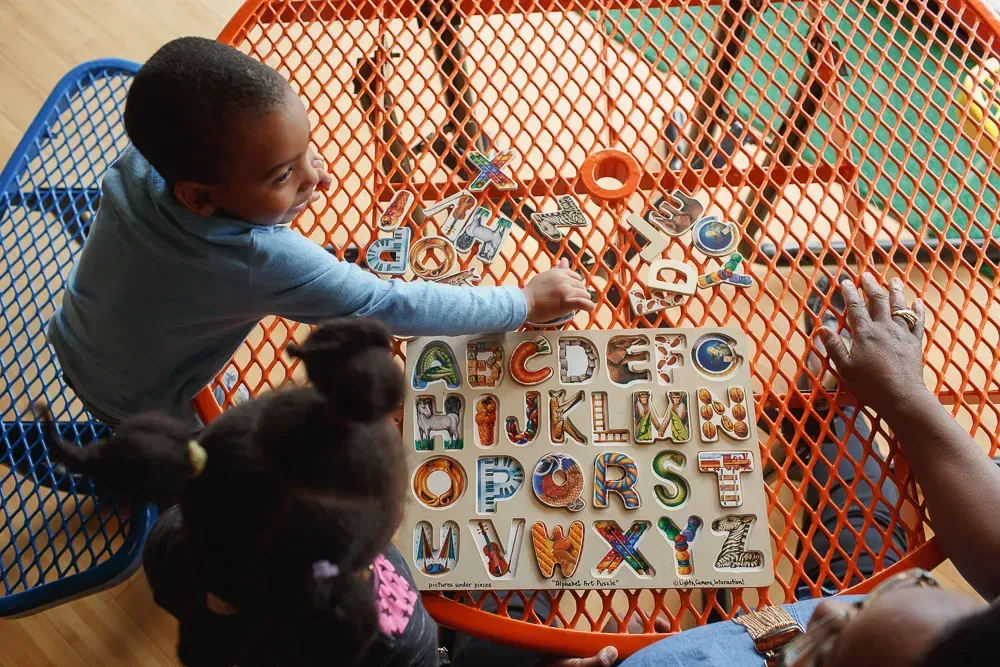 Two children playing with an alphabet puzzle on an orange, mesh table.