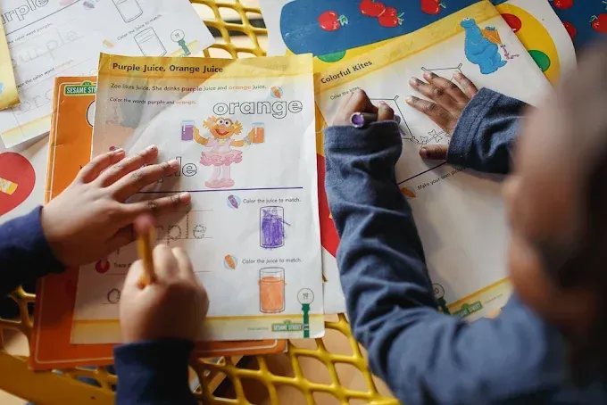 Children at a table create art with guidance from adults. Bright, colorful room.