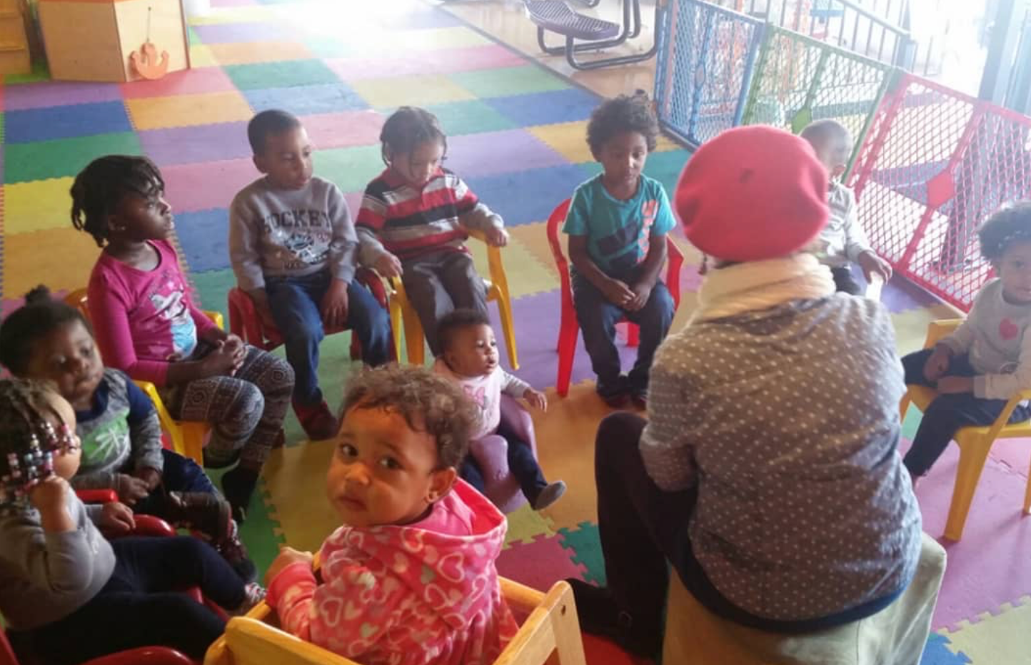 Children sitting in a circle listening to an adult. Brightly colored room, red and blue chairs.