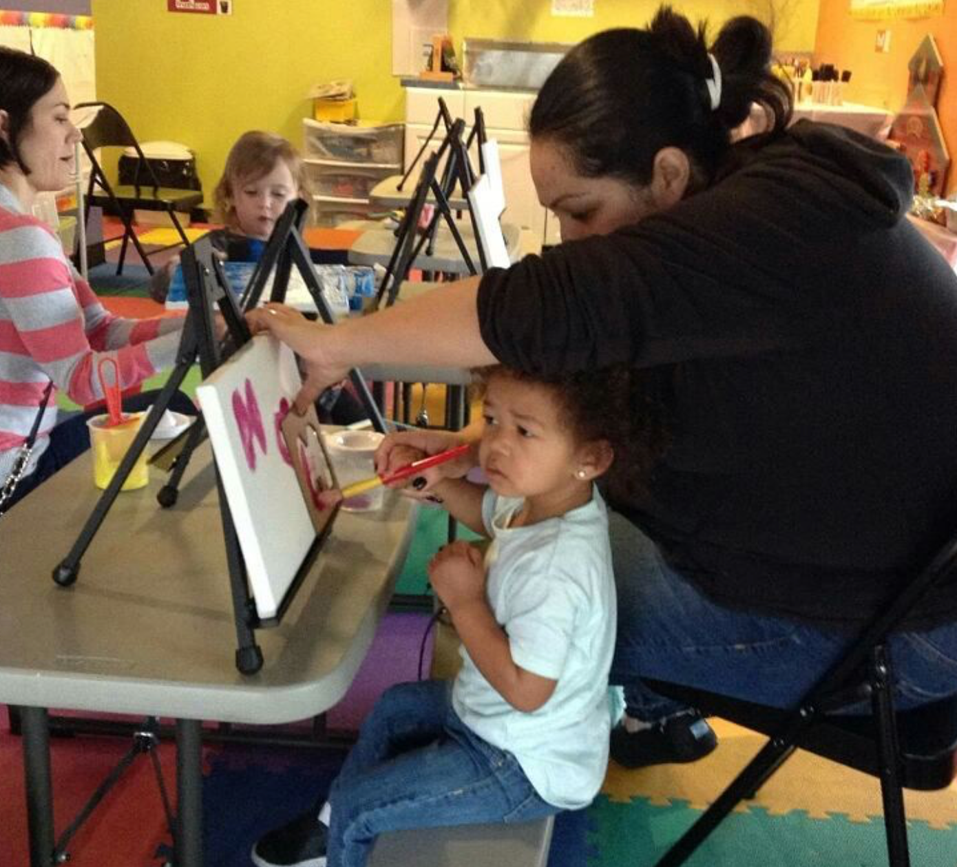A child and adult painting at easels in a colorful room.