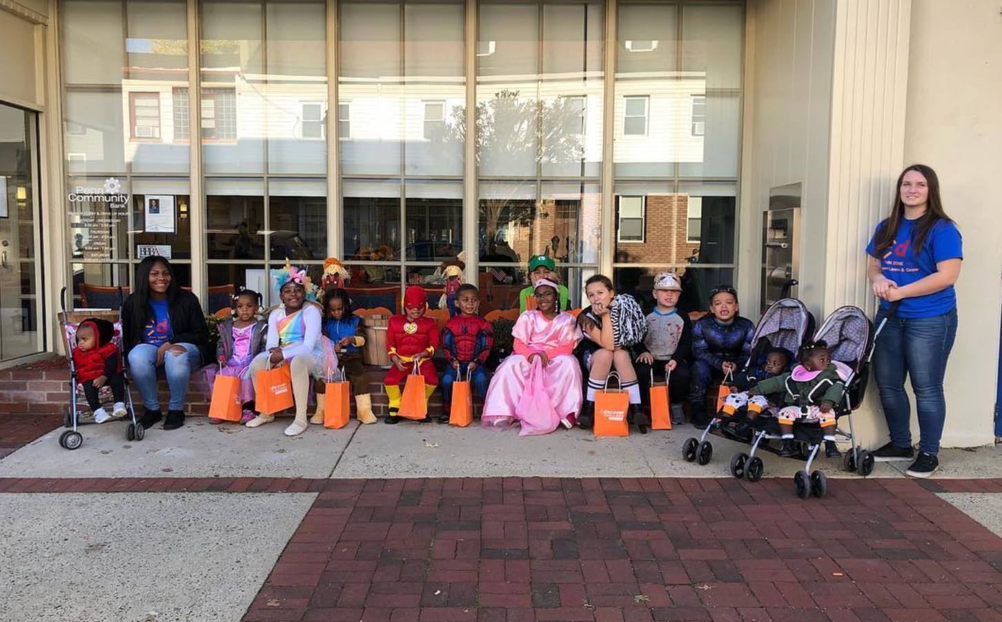 Children in costumes with orange treat bags outside a building; an adult stands nearby.