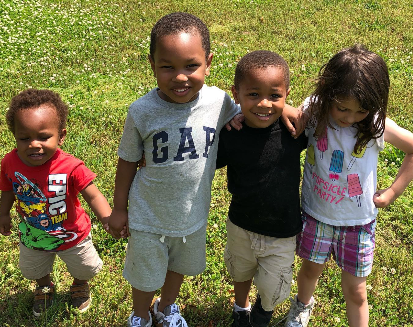 Four children smiling, posing together on grass. Two children have their arms around each other.