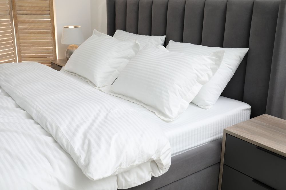 Neatly made bed with white bedding and gray upholstered headboard in a bright bedroom