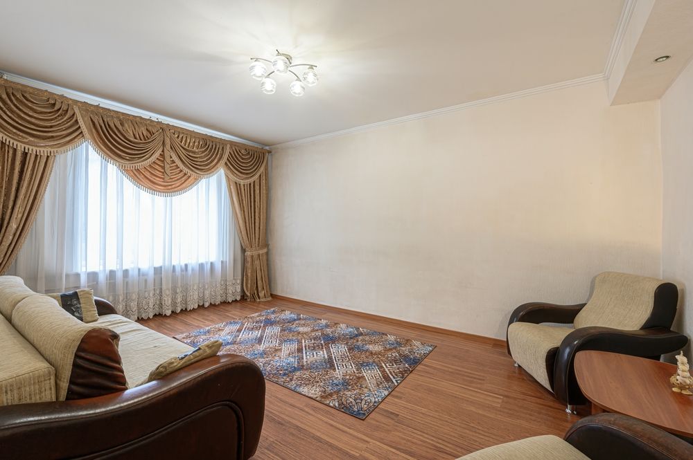 Empty living room with beige walls, patterned curtains, sofa, armchair, rug, and ceiling light