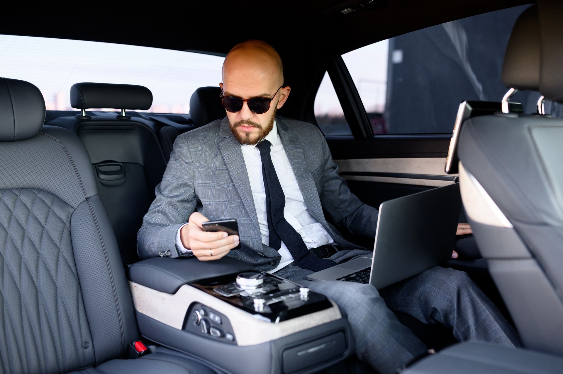 Man in a gray suit and sunglasses using a smartphone in the back seat of a luxury car