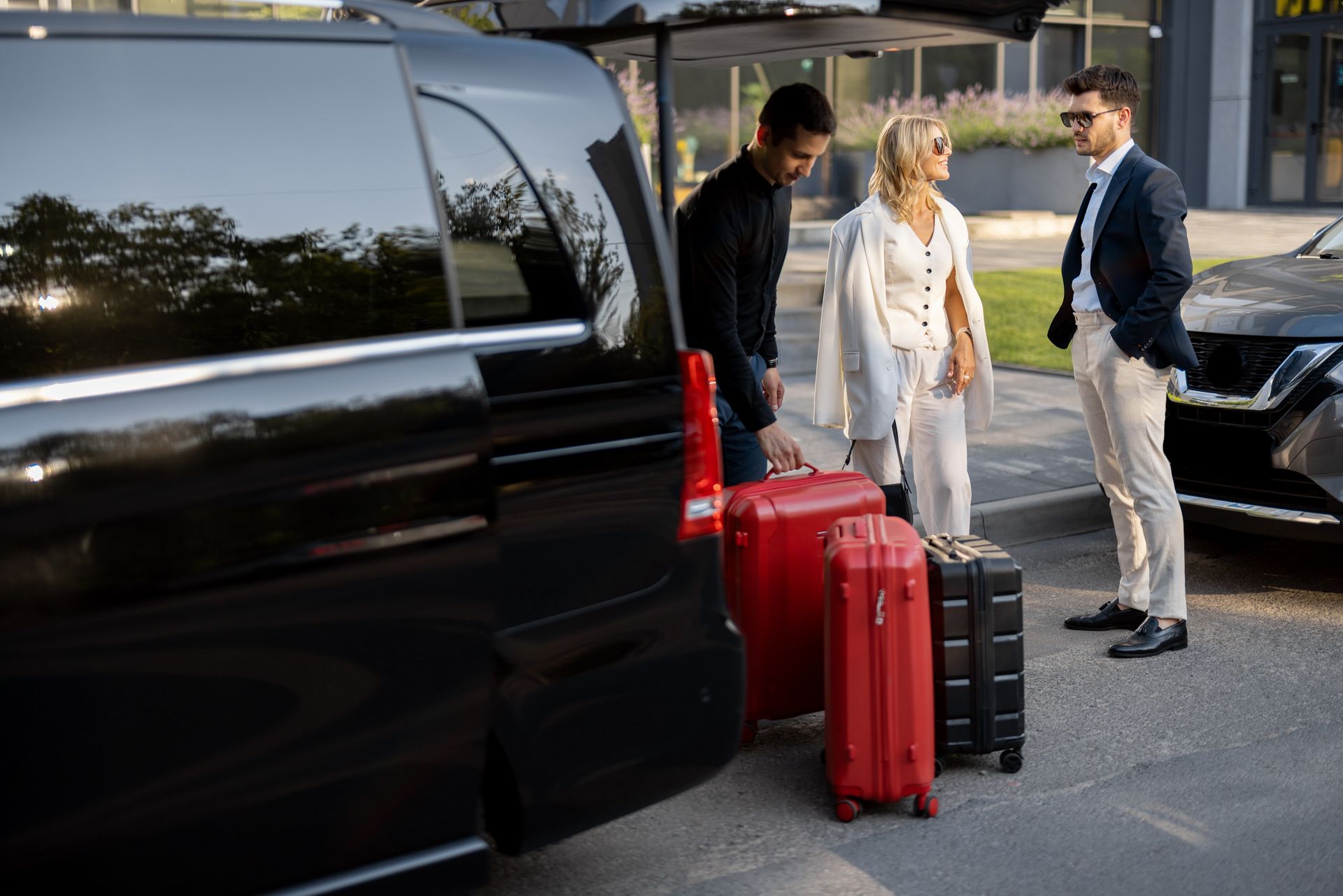 People standing by a black SUV and luggage, including a red suitcase, on a driveway