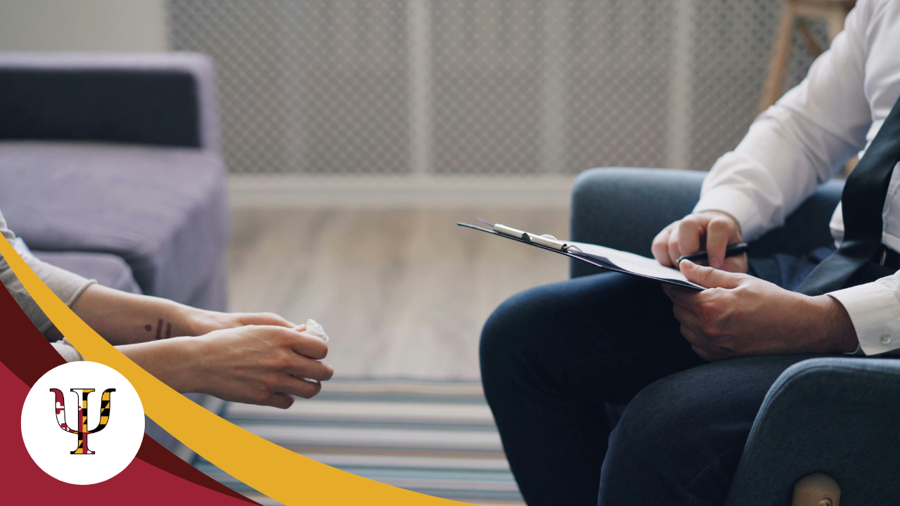 Two people seated in a meeting, one holding a notebook and pen, with a couch and table in the background.