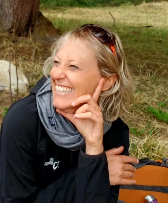 Donna sorridente, all'aperto, con giacca scura, sciarpa, occhiali da sole in testa, mano vicino al viso, borsa arancione.