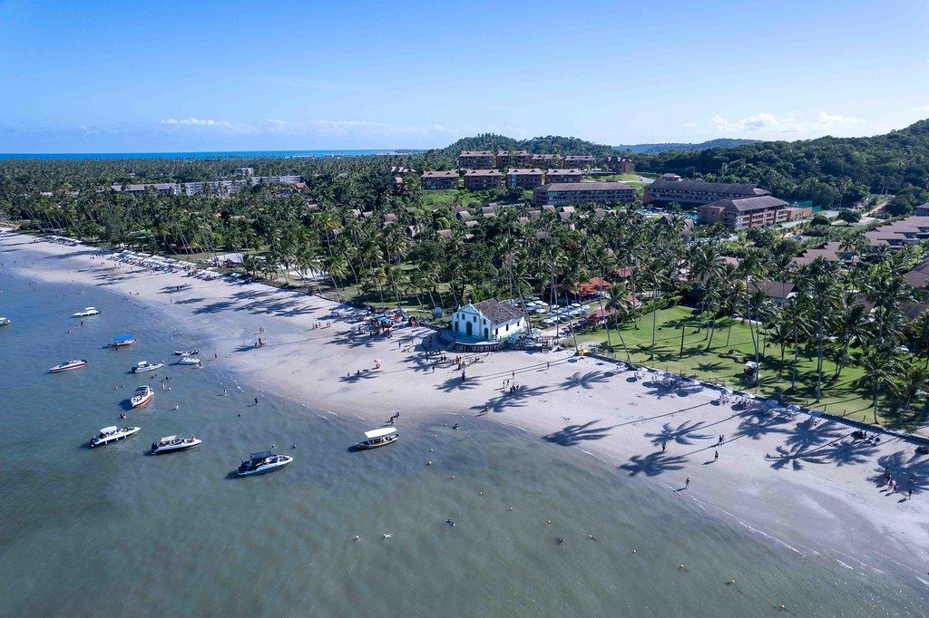 Vista aérea de uma praia de areia branca com barcos e palmeiras. Um pequeno edifício branco está situado na costa.