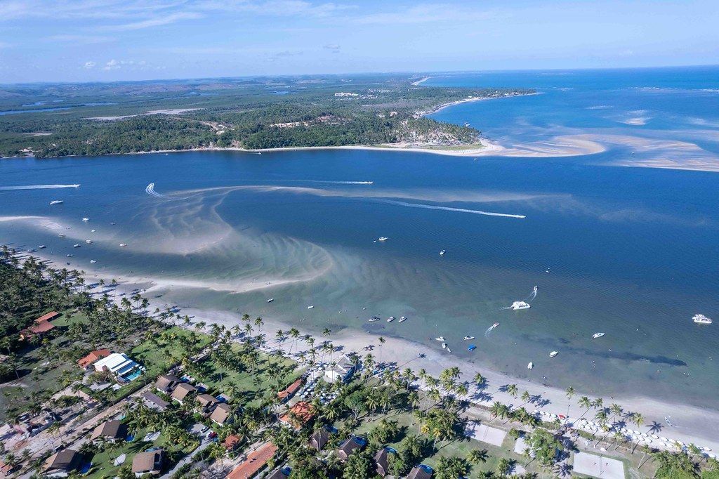 Vista aérea de uma praia com areia branca, água turquesa e uma costa verdejante. Barcos e edifícios pontilham a paisagem.