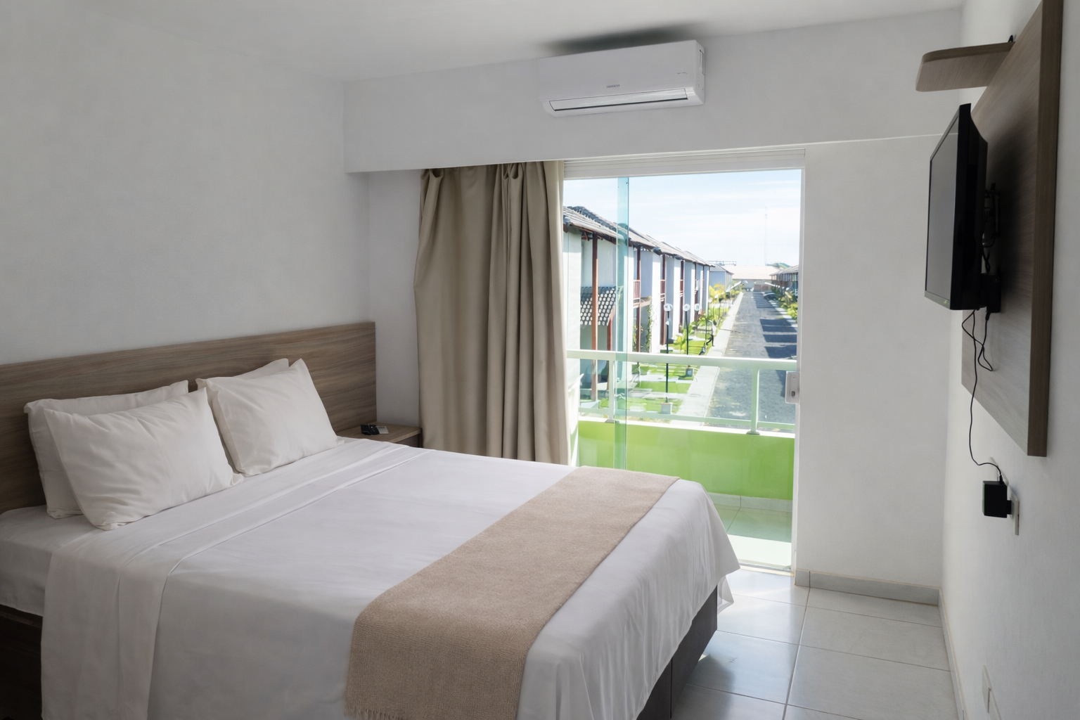 Quarto de hotel com decoração em tons de branco e bege, duas camas, uma pequena TV e uma varanda com vegetação.