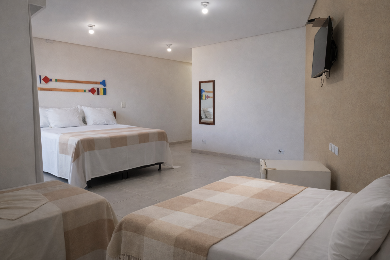Quarto de hotel com três camas: duas de casal e uma de solteiro, em tons neutros. TV na parede.