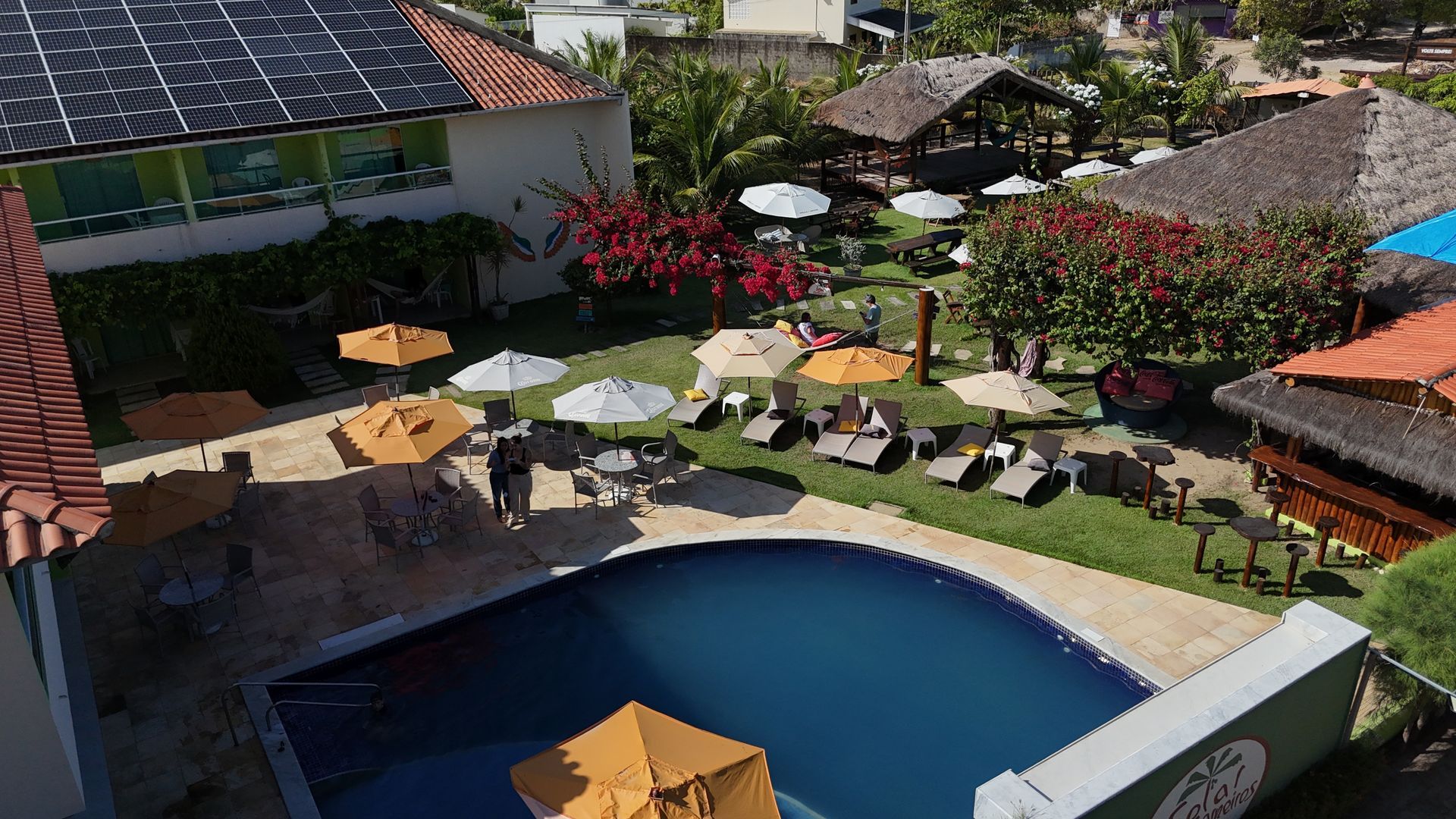 Área da piscina com espreguiçadeiras, guarda-sóis e estruturas com telhado de palha. Gramado verde, árvores e construções.