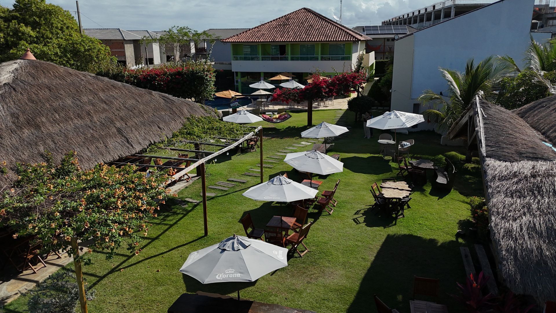 Restaurante ao ar livre com estrutura de telhado de palha, mesas e guarda-sóis sobre grama verde, e um prédio de dois andares ao fundo.