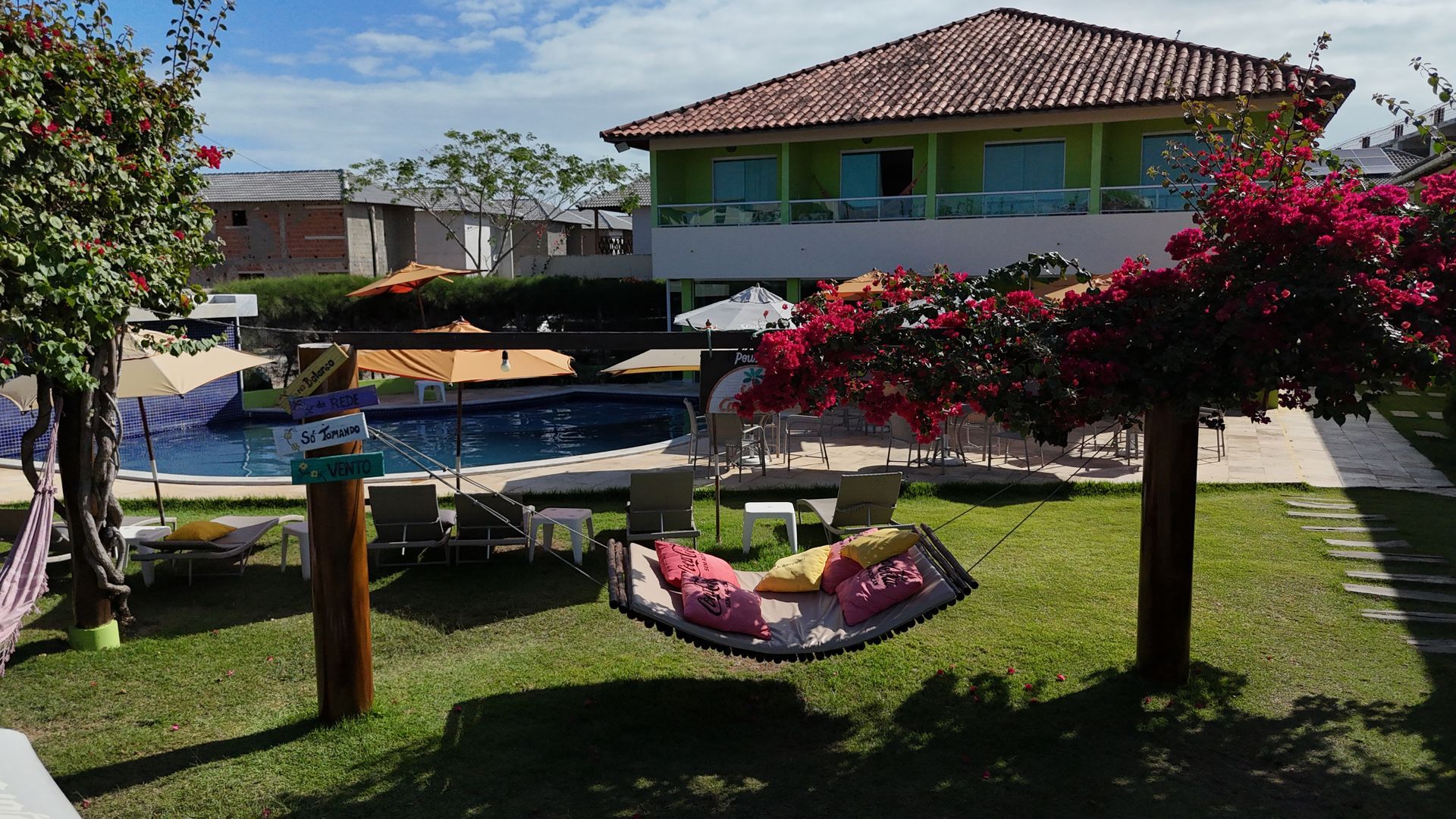 Quintal verdejante com rede, piscina e, ao fundo, um prédio de dois andares com telhado de telha vermelha.