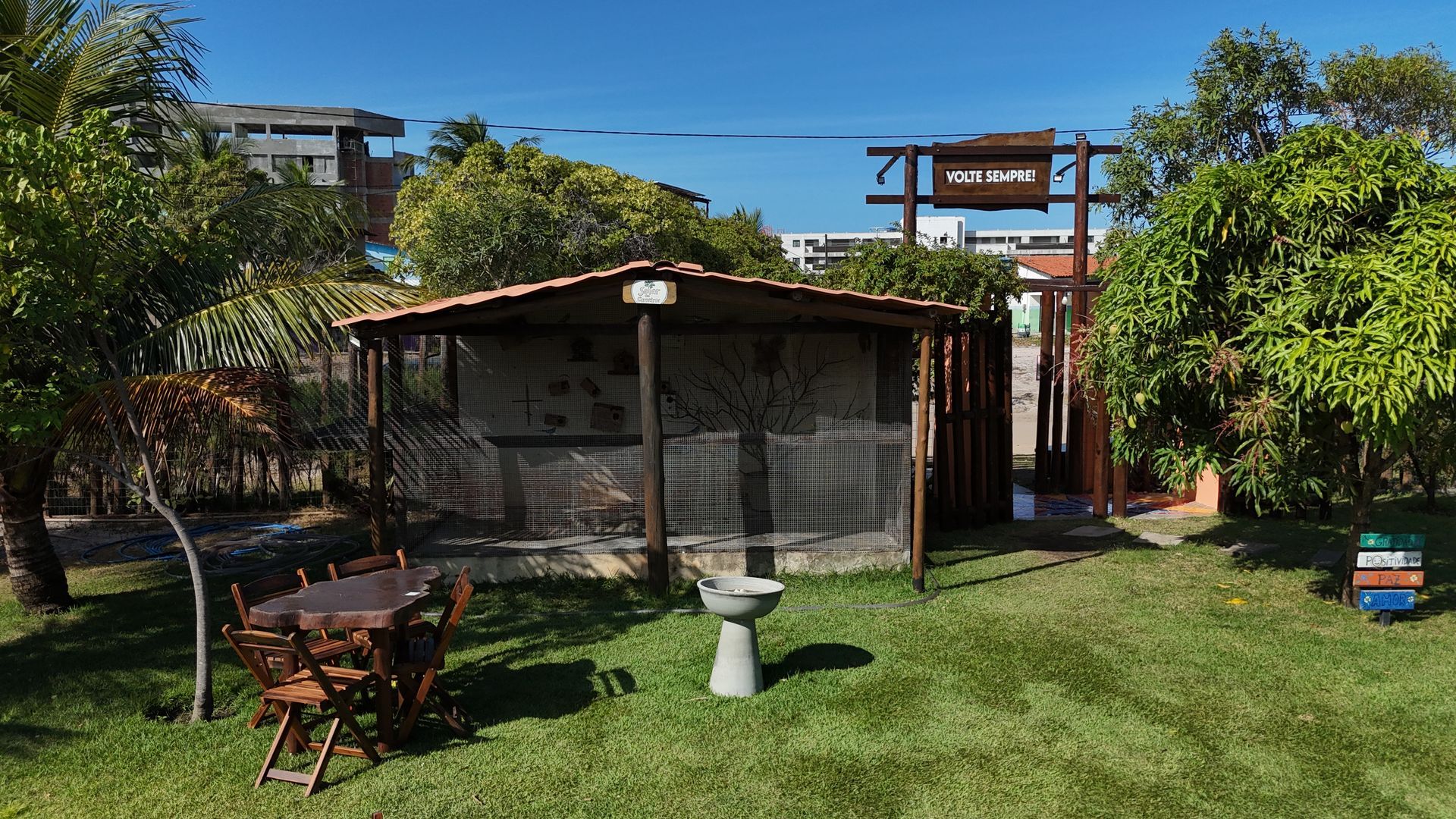 Gazebo de madeira, bebedouro para pássaros e mesa em um gramado. Portão de entrada com placa, cercado por árvores. Dia ensolarado.