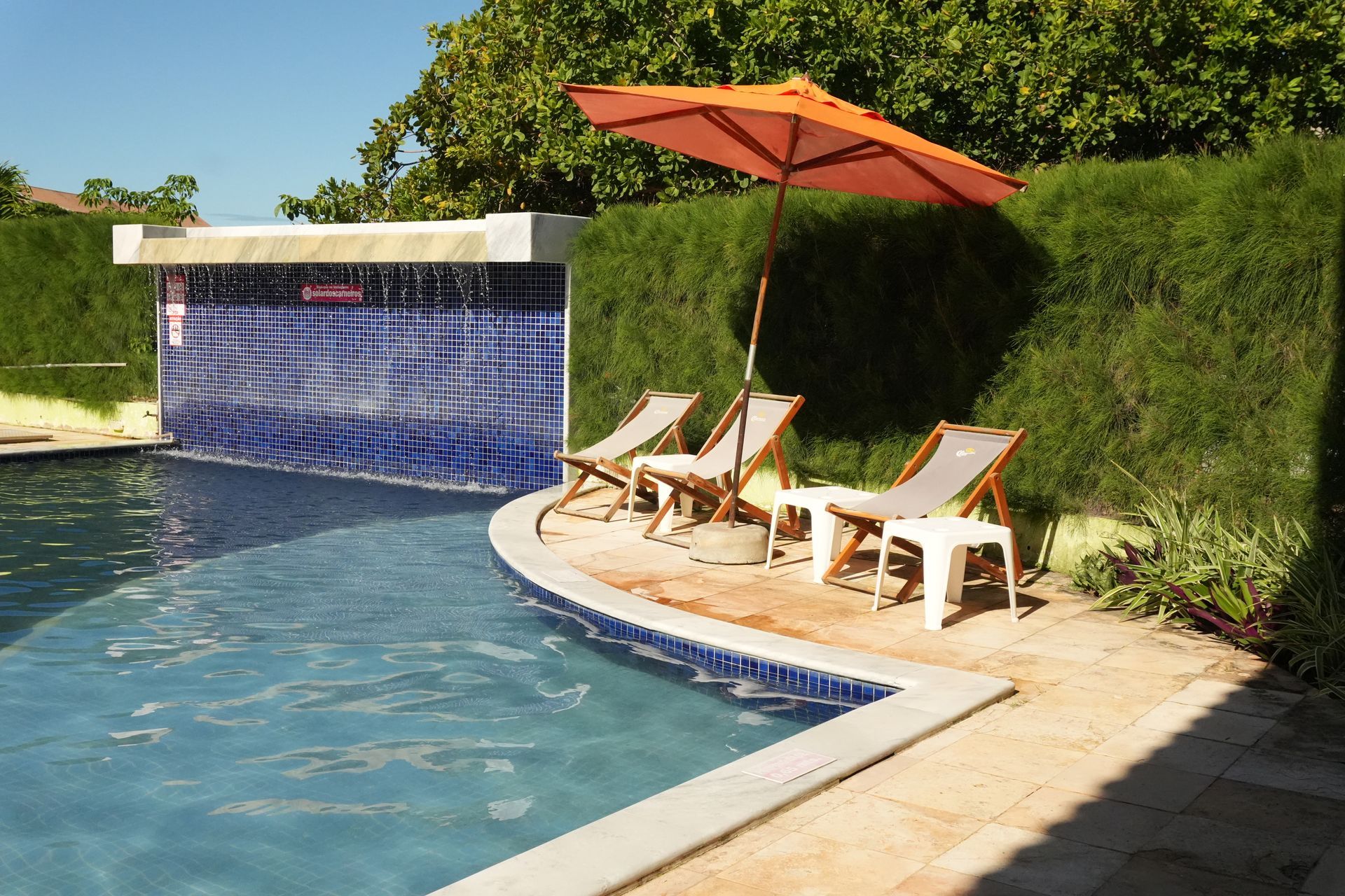 Cena à beira da piscina com uma cascata revestida de azulejos azuis, espreguiçadeiras sob um guarda-sol laranja e uma sebe verde ao fundo.