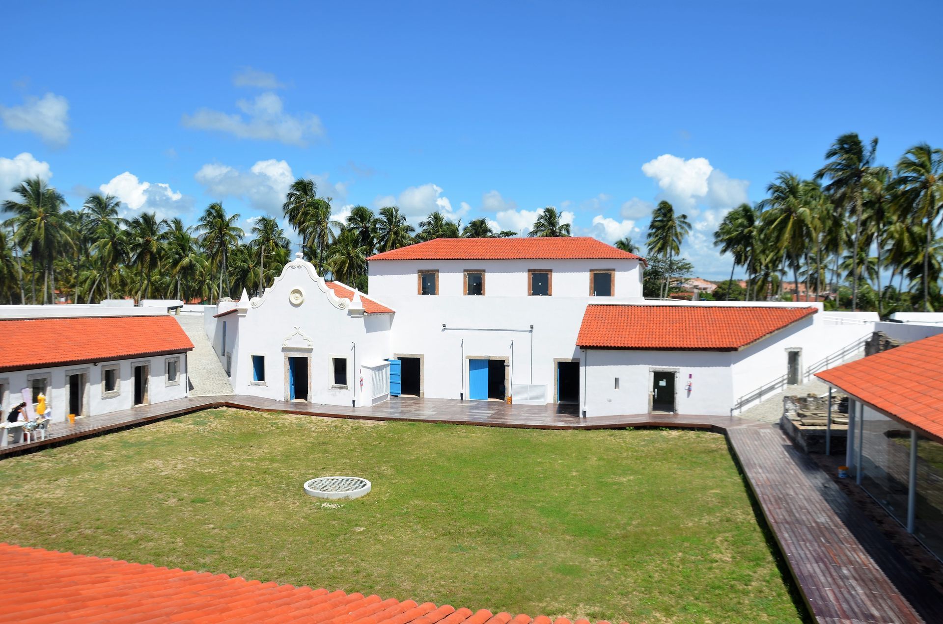 Complexo de edifícios brancos com telhados de telha vermelha, portas azuis, gramado verde e palmeiras sob um céu azul.