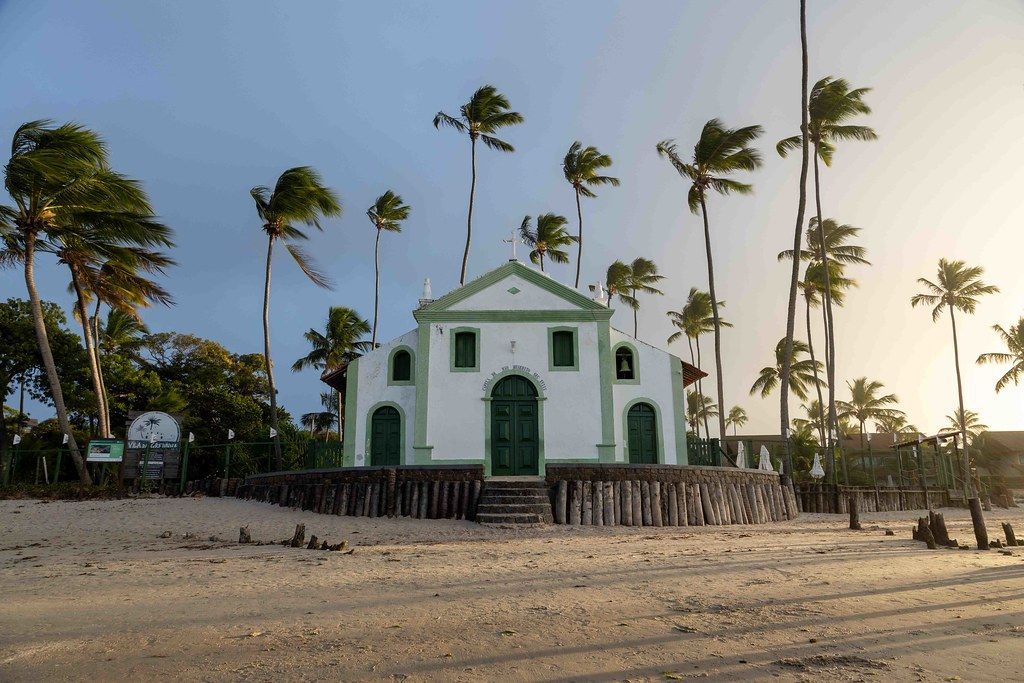 Igreja branca com detalhes verdes em uma praia, rodeada por palmeiras sob um céu crepuscular.